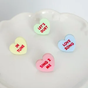 Four pastel candy heart stud earrings with music-themed phrases on a white dish.